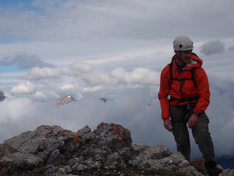 Jon smiling on a ridge above the clouds near Jasper, helmet on, mountains breaking through the mist behind him