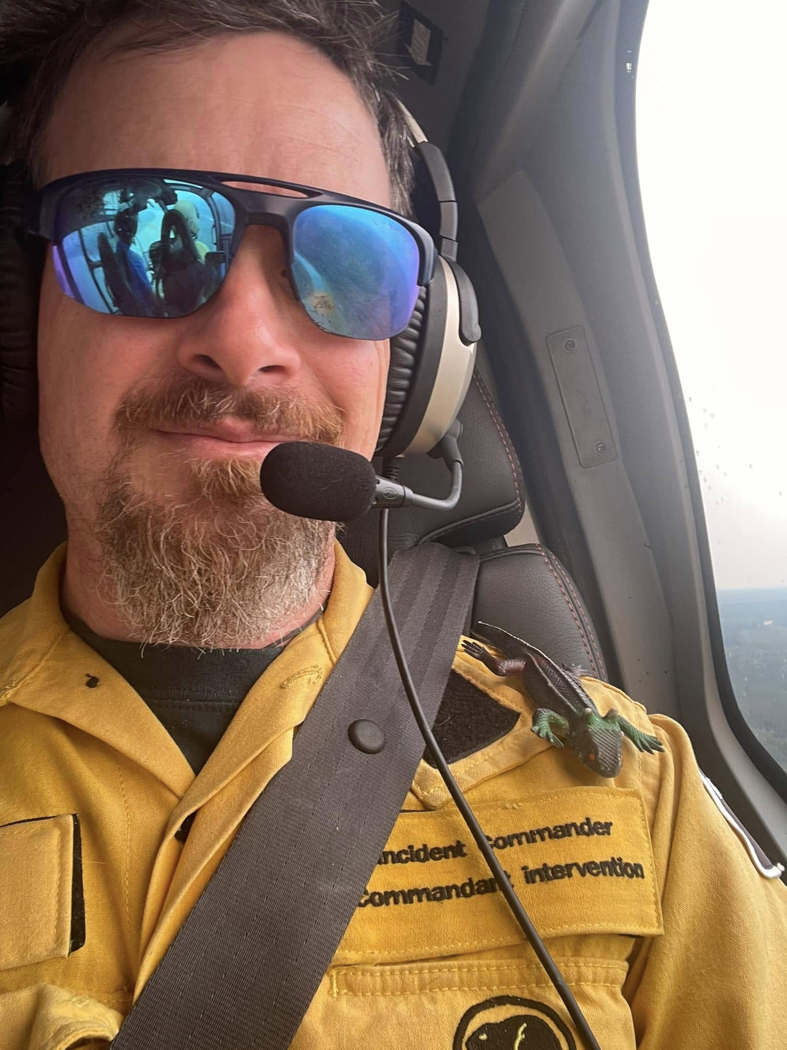 Jon wearing an Incident Commander jacket and headset inside a helicopter, with Lizzy the toy lizard riding on his shoulder