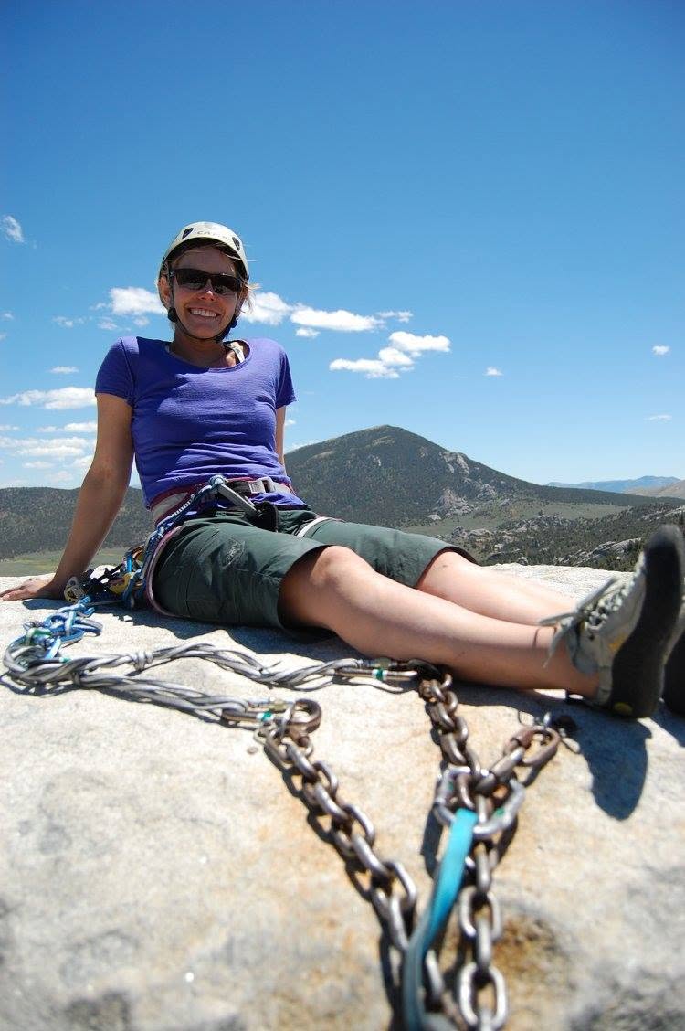 Rudie smiling at a belay anchor with chains in the foreground and mountains behind